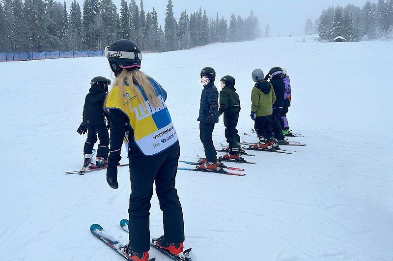 En ledare Alla på snö står tillsammans med några barn i en skidbacke, de har skidor och hjälm.