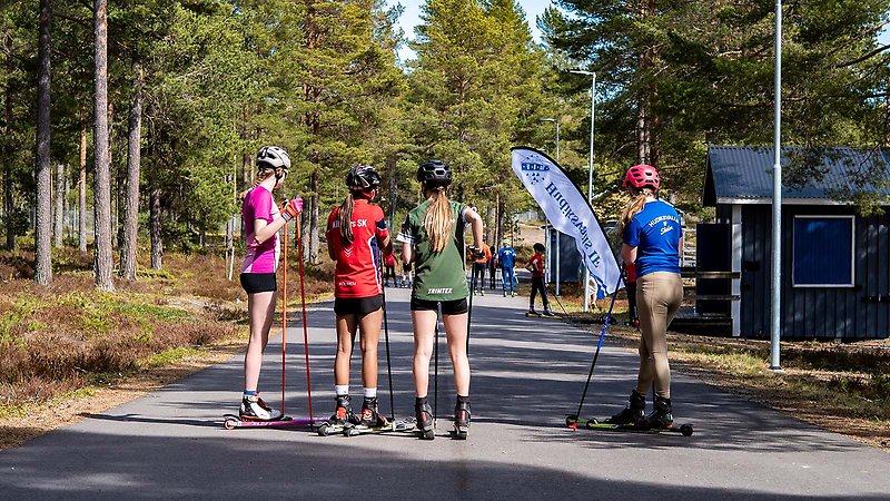Fyra ungdomar på rullskidor står med ryggen till på en asfalterad väg i en skog. De bär olika klubbtröjor i olika färger.