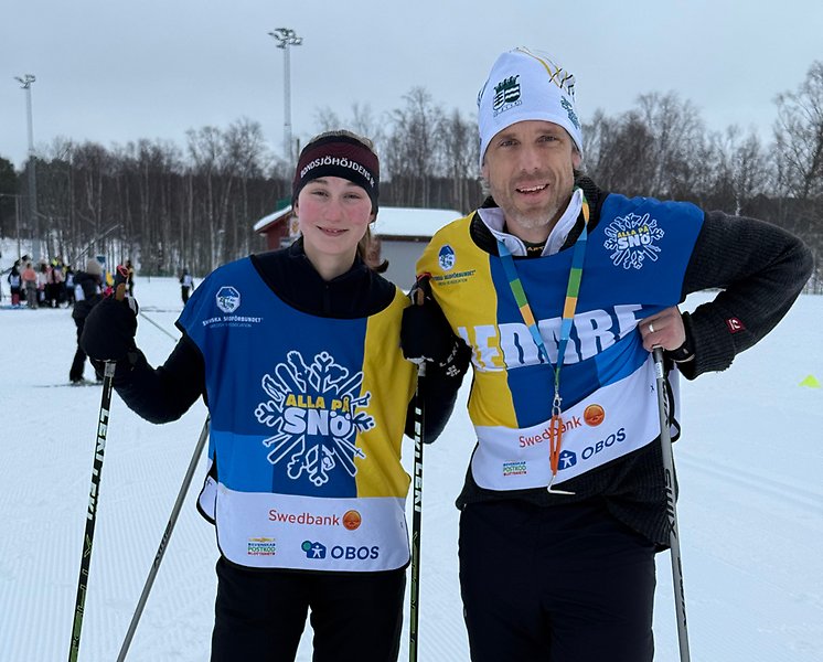 Två glada ledare i Alla på snö-västar står i snön på längdskidor